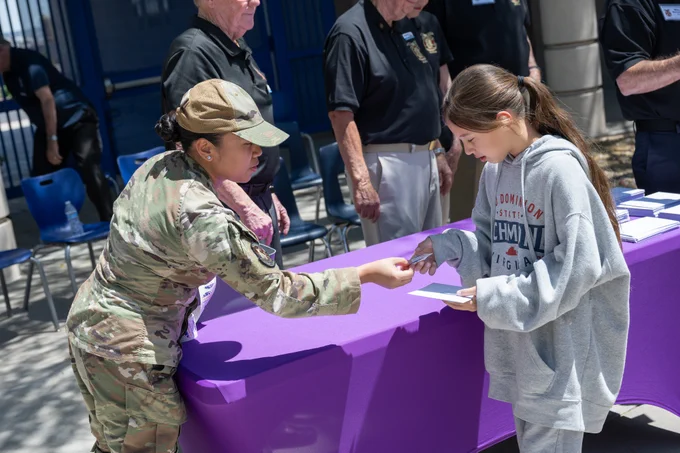 A military personnel in uniform hands a document to a young woman wearing a gray hoodie at an outdoor event with a purple tablecloth.