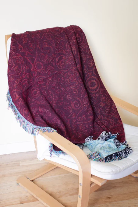 A blanket in dark red, with the AO3 and OTW logo woven into it as a pattern. It is draped over a chair.
