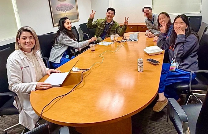 A group of people is seated around an oval conference table, engaging cheerfully. One person holds a laptop. Others are gesturing playfully with peace signs. There are drinks and snacks on the table. A framed painting is visible on the wall.