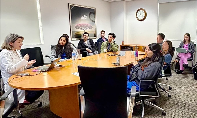 A group of people sit around a large conference table in an office setting. One person at the head of the table gestures while speaking, and others listen attentively. A laptop, water bottles, and notebooks are on the table. A framed nature picture is on the wall, and a clock is visible.