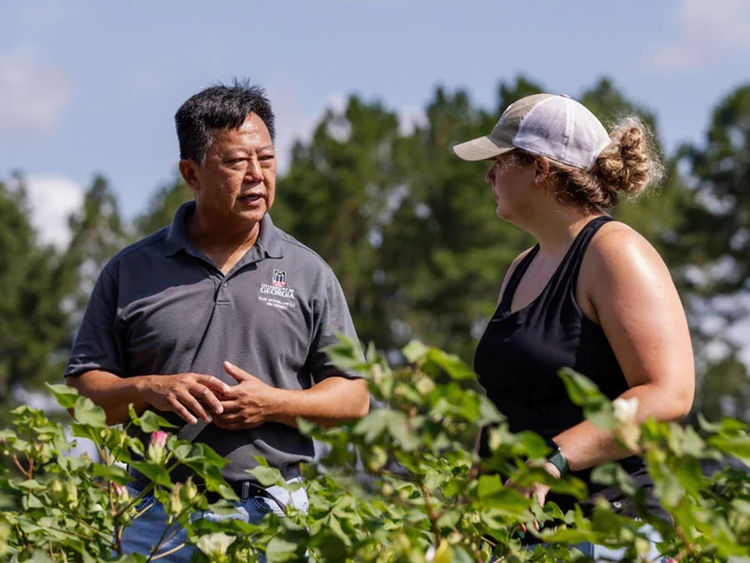 Two people are having a conversation in a lush, green outdoor setting. One individual is wearing a polo shirt, while the other is dressed in a tank top and cap. Trees are visible in the background under a blue sky.