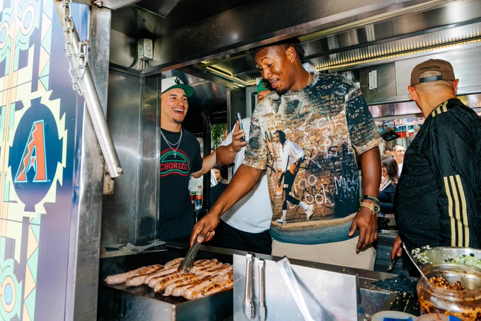 Alek Thomas laughs as Geraldo Perdomo cooks hot dogs in a food truck. 