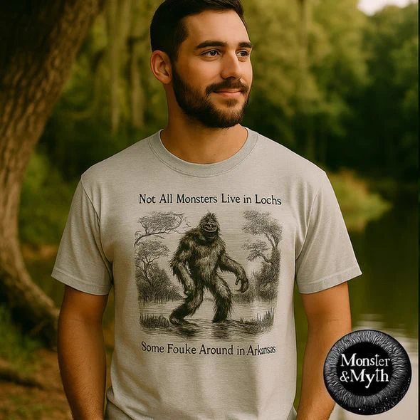 A man stands near a wooded swamp in Arkansas wearing a light gray T-shirt. The shirt features a vintage-style sketch of the Fouke Monster, a hairy Bigfoot-like creature walking through a swamp. Above the image, it reads “Not All Monsters Live in Lochs,” and below, “Some Fouke Around in Arkansas.” The Monster & Myth logo is displayed in the corner.