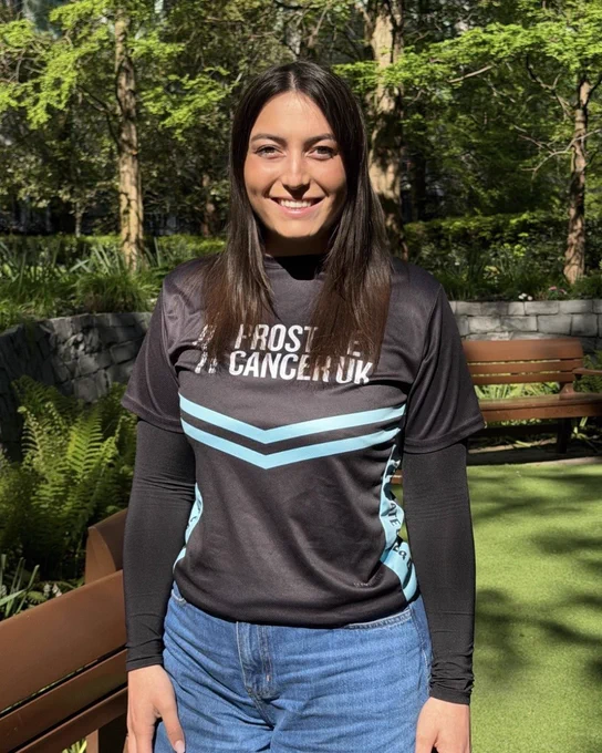 Annie is smiling while standing outdoors in a garden. She is wearing a black Prostate Cancer UK running t-shirt and blue jeans. Surrounding her are green plants and trees, and she is standing on a grass or turf surface next to a wooden bench.