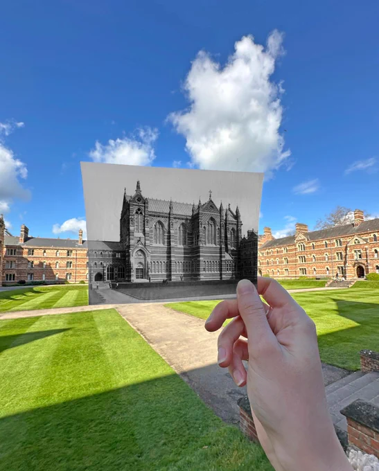 A hand holds a black-and-white historic photo of Keble Chapel aligned with the present-day college quad, showing red-brick buildings, green lawn, and blue sky.
