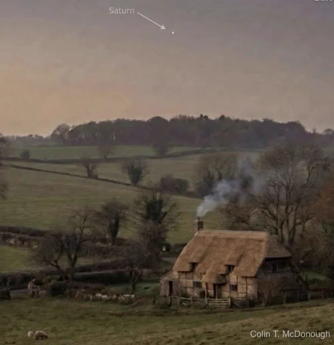 A peaceful twilight photograph of the English countryside featuring a historic timber-framed thatched cottage with smoke rising from the chimney. In the soft, hazy sky above the rolling green hills, a faint dot is identified by a subtle arrow and label as the planet Saturn. The name "Colin T. McDonough" is visible in the bottom right corner. The image illustrates the Roman and Anglo-Saxon origins of the name "Saturday."
