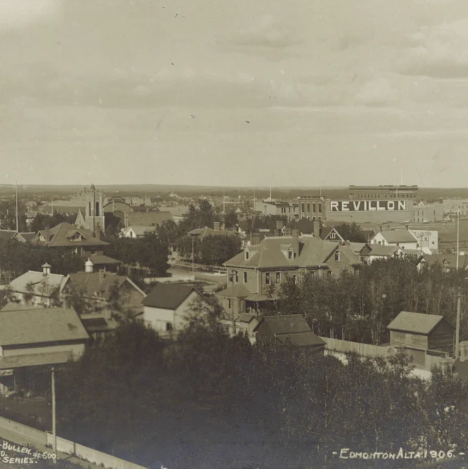Wide panoramic photograph of a town with numerous small houses, tree-lined streets, and a mix of residential and larger institutional buildings. In the mid-distance, a large commercial building displays the word REVILLON painted prominently across its upper facade. Several church spires and taller structures rise above the rooftops. The foreground shows fenced yards, utility poles, and clusters of trees. Along the bottom edge of the image, faint text reads Sanderson-Bullen Photo, Empire Series and Edmonton Alta 1906.
