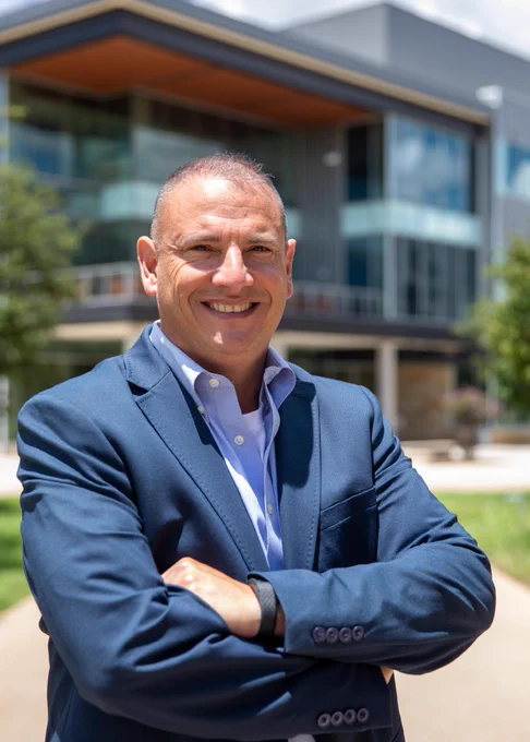 A man stands in front of a building that has glass windows. He is wearing a navy blue suite and has his arms crossed in front of him.