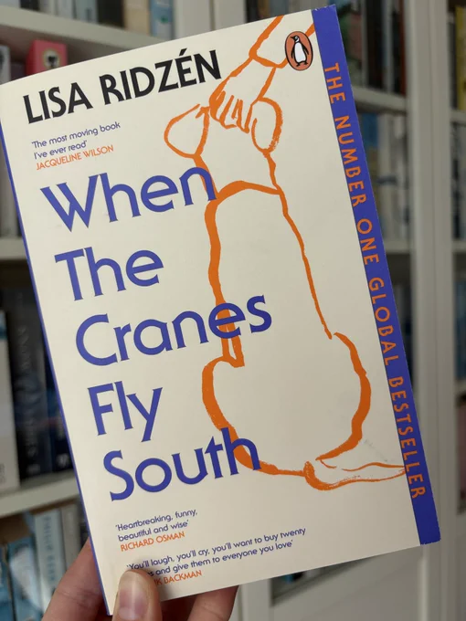 A book is held up in front of bookshelves. The book is called ‘When The Cranes Fly South’ by Lisa Ridzén. The cover is a pale cream, with the outline of a sitting dog with a hand on its head drawn in orange on the right side. The title, in blue, is on the left of the cover.