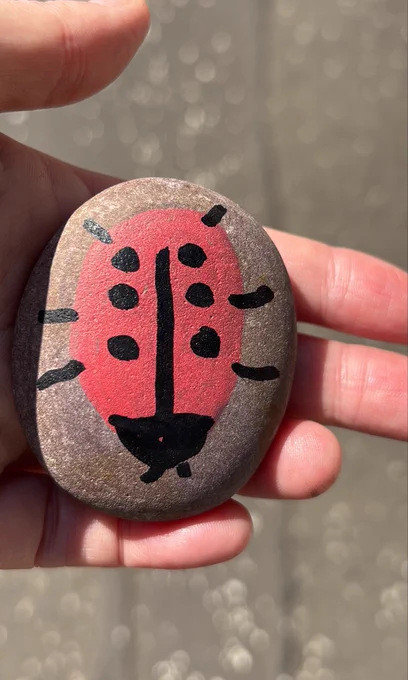 A hand holds a smooth oval stone painted with a simple red and black ladybird. The background is an outdoor surface in sunlight