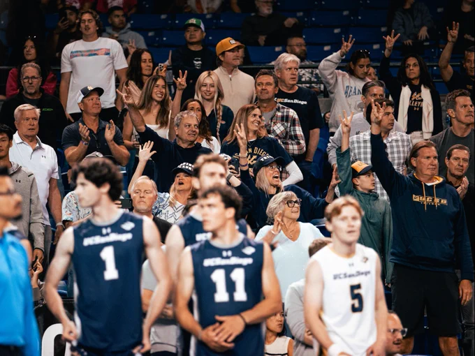 A crowd of enthusiastic fans in a sports arena, many wearing "UC San Diego" apparel. They display excitement, cheering and making hand gestures, with some making Trident signs. In the foreground, four athletes in "UC San Diego" uniforms stand in front of the crowd.