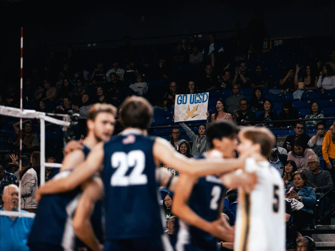 A group of volleyball players in navy and white UC San Diego uniforms are gathered on the court. In the background, spectators in the stands hold a sign reading "Go UCSD!"