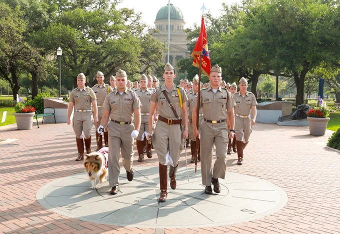 A group of people in uniform marches in formation on a brick path, holding flags. Reveille X walks alongside them. In the background, trees are visible, and a domed building is partially seen.