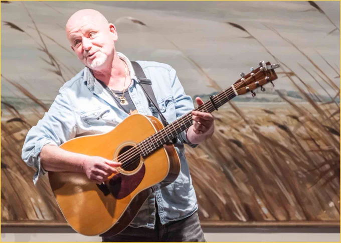 Photo of Anthony John Clarke standing, singing and playing guitar. AJ is wearing a black t-shirt beneath a pale blue shirt, and dark jeans.