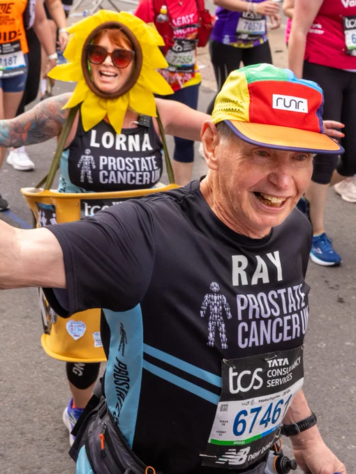 A runner called Ray wearing a colourful hat and Prostate Cancer UK shirt is running and smiling. Another person behind, Lorna, is wearing a sunflower headpiece, also wearing a Prostate Cancer UK shirt. Both are running in the London Marathon.