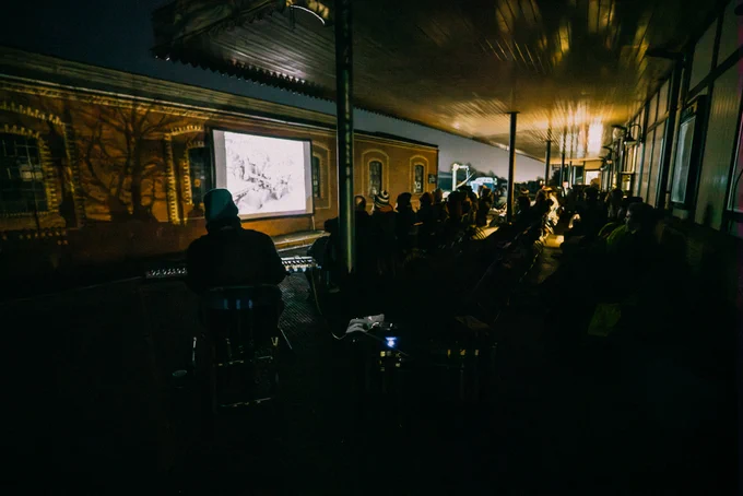 A wide shot of the illuminated film screen and the audience on the train platform.