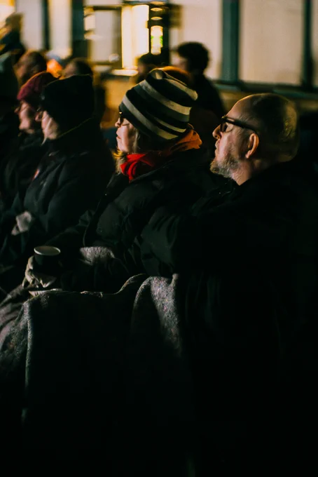 Dimly lit audience members sit on the train platform.