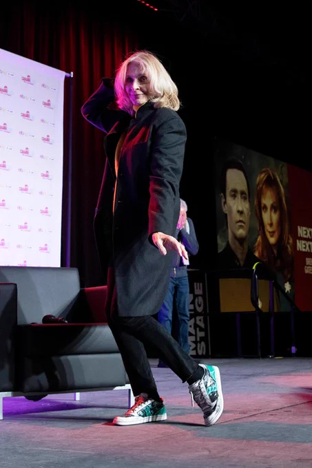 Gates McFadden in black jeans and black coat with a green jumper under and shoesbyjax Beverly Crusher custom sneakers. She's posing for photos with a hand behind her head and the other delicate on the side, a little bit like dancing.