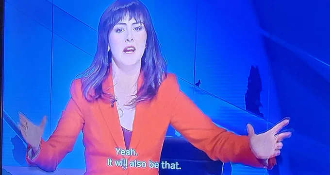 The image shows a woman with dark hair and bangs captured in a medium shot from a television screen, where she is dressed in a vibrant orange blazer over a dark top. She is gesturing expansively with both hands while speaking, set against a modern, minimalist studio backdrop characterized by cool blue tones and sharp geometric lines. Subtitles at the bottom of the frame read, "Yeah. It will also be that," indicating she is in the middle of a discussion or interview. The photograph retains the slight pixelation and blue-tinted glow typical of a picture taken directly from a digital monitor, emphasizing its origin as a broadcast still.