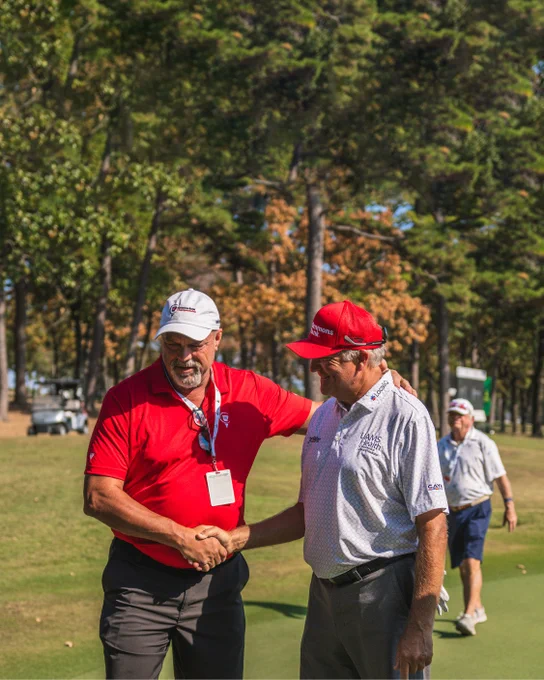 Two people are shaking hands on a golf course. Both are wearing golf attire, including caps with logos. The course is surrounded by trees, and two golf carts are visible in the background. One person is wearing a red shirt and lanyard.