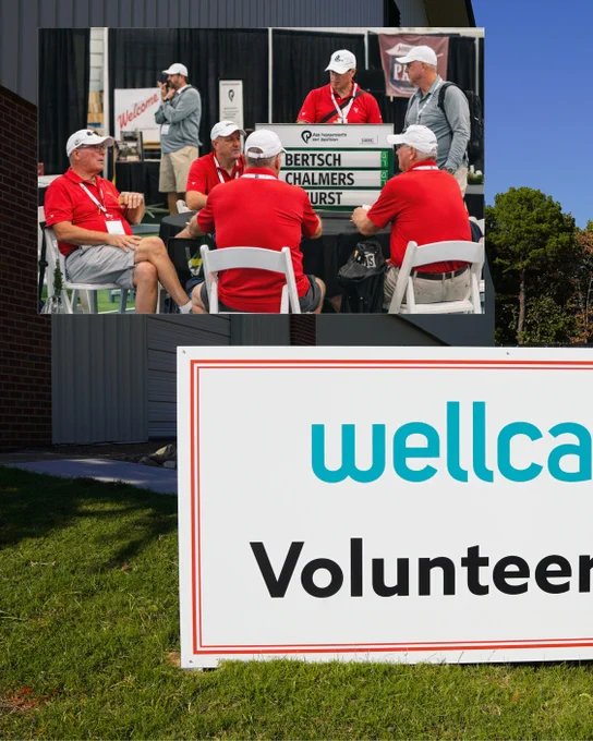 A group of individuals wearing red shirts and white hats sit around a table with a sign displaying names. Nearby, a large "Wellcare Volunteers" sign is visible on a grassy area.