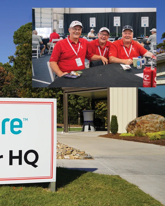 A group of smiling people wearing matching red shirts with badges seated around a table indoors. In the background, more people are visible in a large room. The lower part of the image shows a large sign with partially visible text and the exterior of a large building surrounded by trees and landscaping.
