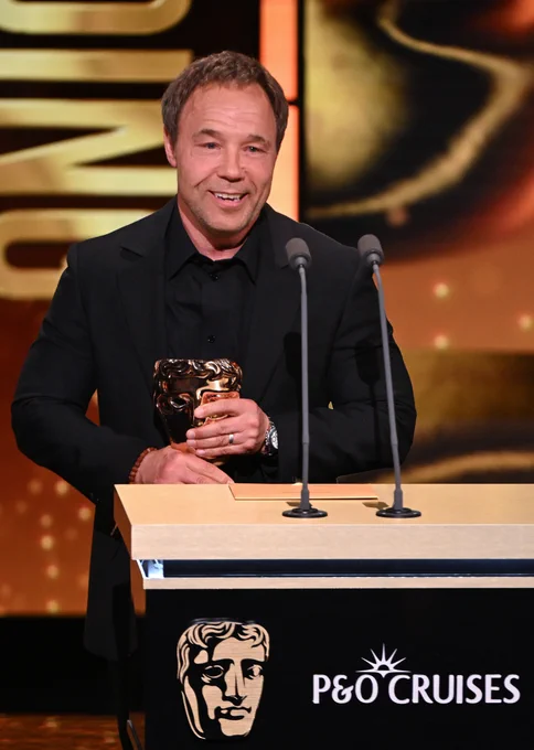 Stephen Graham in a black suit holds a BAFTA award, standing at a podium. Background displays a warm, golden hue. He appears excited and proud.