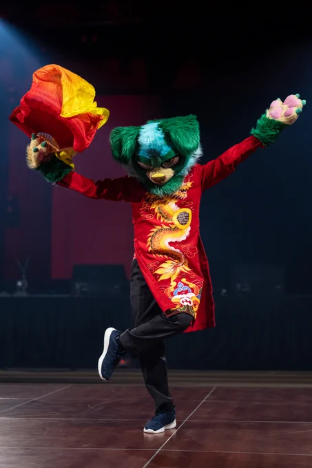 A performer in a colorful fursuit stands poised on stage, wearing a vibrant red outfit with a dragon design. The scene is lively and dynamic.