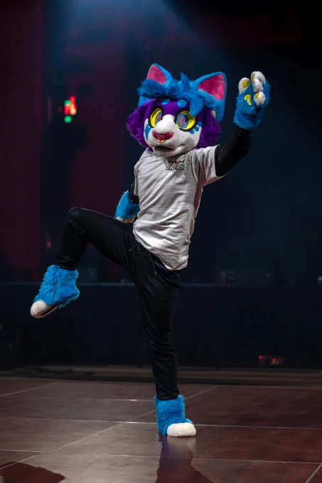 A performer in a vibrant fursuit expresses a joyous dance pose on stage. The setting suggests a lively, creative atmosphere.