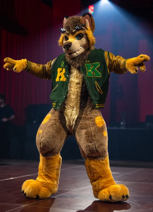 A person in a furry costume poses on stage, wearing a green and yellow letterman jacket. The setting appears to be a lively event with a spotlight highlighting the character.