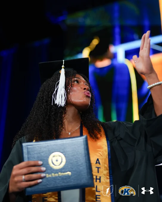 A graduate in cap and gown holds a diploma cover from Kent State University, standing on a stage with banners and logos including Under Armour.