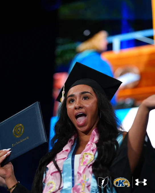 A person in graduation attire is celebrating, holding a diploma. The robe features a Kent State University logo. The background shows a large display screen with another individual in academic regalia. Logos for Kent State University and Under Armour are visible.