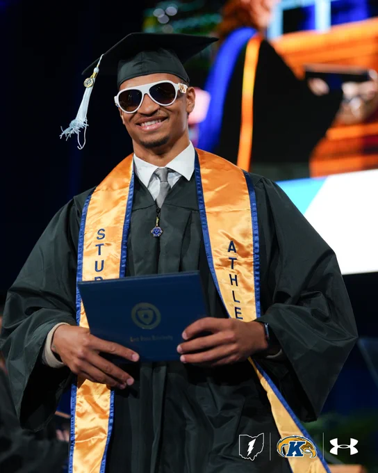 A person wearing a graduation cap, gown, and stole, holds a diploma folder while smiling. They wear large sunglasses and the stoles read "Student Athlete" with Kent State University logos visible at the bottom.