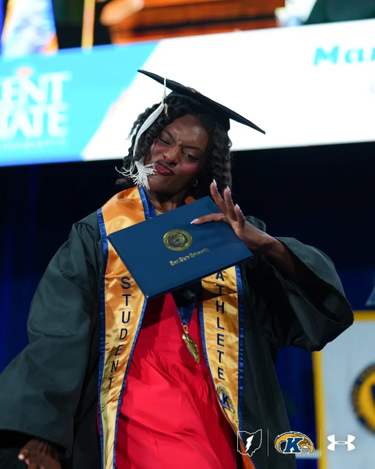A graduate in a red outfit holds a diploma while on stage. They wear a cap and gown with "Student Athlete" sashes, and the background displays Kent State University insignia and logos.