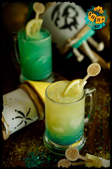 Promotional photograph of the official SoTFest alcoholic drink, “Ghostly Grog”, served in clear glass tankards filled with ice. The foreground drink has a glowing layered appearance, fading from bright turquoise blue at the bottom to pale golden yellow at the top, garnished with a lemon slice and a branded wooden stirrer featuring the SoTFest logo. A second bright green version of the drink appears blurred in the background. The drinks sit on a dark wooden table scattered with turquoise and gold glitter confetti, alongside tipped-over pirate-style tankards. The SoTFest 2026 logo is displayed in the top right corner, giving the image a festive pirate-themed atmosphere.