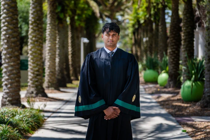 Yeran Gamage in a graduation gown standing on a tree-lined sidewalk. The setting is sunny, with palm trees and large green planters visible in the background.