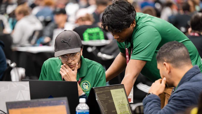 A group of people gathered around laptops at a tech event. One person is wearing a Puma cap and a green shirt with a circular emblem. The setting appears busy, with many individuals in the background working on computers.