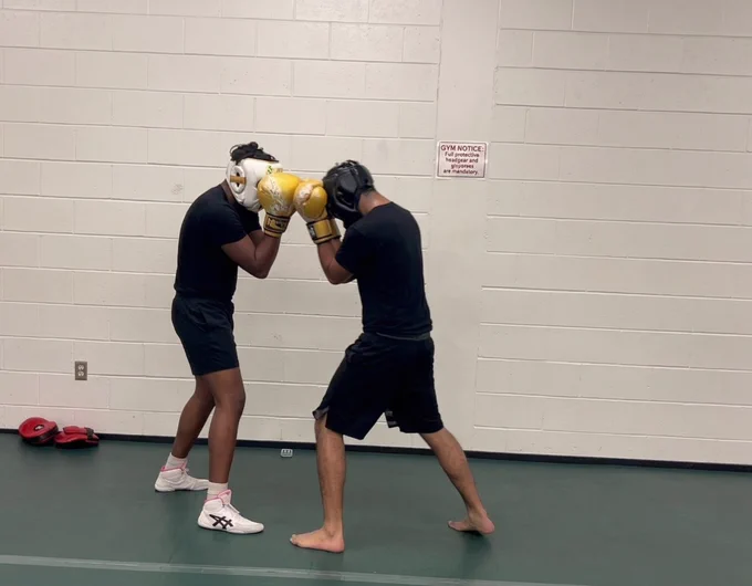 Two people are boxing in a gym. Both are wearing protective headgear and boxing gloves. One person has golden gloves, while the other's are white. They are positioned in front of a white brick wall, with a small sign visible in the background. Red focus pads are on the floor nearby.