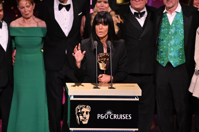 Claudia Winkleman in black speaks at a podium with a golden BAFTA TV Award statue, surrounded by celebrities who competed in formal attire. Emotions are celebratory and formal.