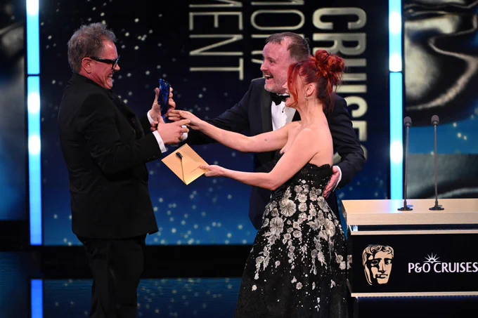 Three people celebrate on stage at an award ceremony. Alan Carr, Chris McCausland and Dianne Buswell. They are smiling as they hold a trophy and envelope. The backdrop features a logo and stars.