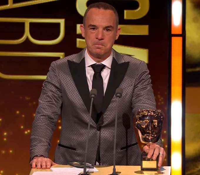 Martin Lewis in a gray suit stands at a podium with dual microphones, holding a BAFTA trophy. The background features golden letters, creating a formal ambiance.