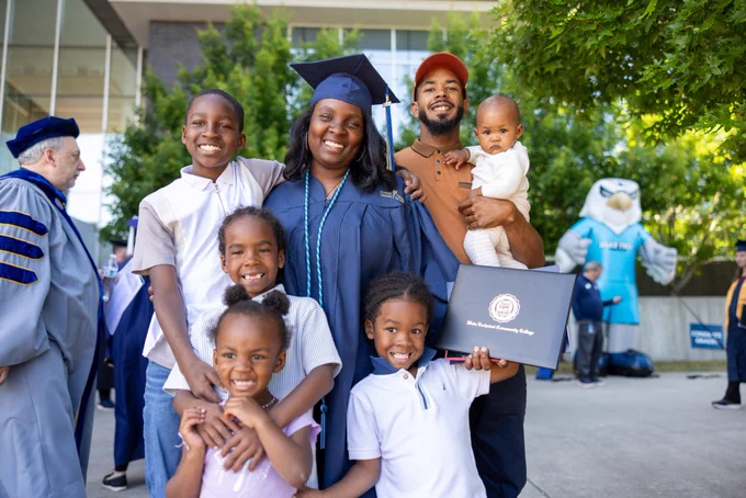 Graduate in a navy cap and gown stands outdoors holding a diploma while surrounded by several children and an adult holding a baby; trees and other attendees are visible in the background.