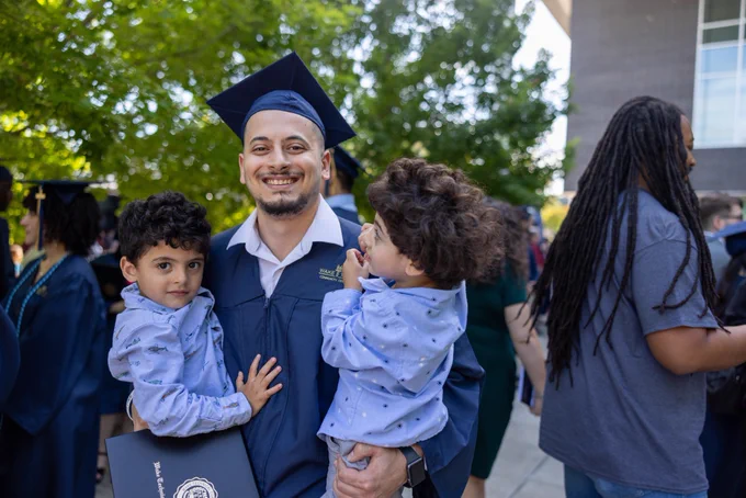 Graduate in a navy cap and gown stands outdoors holding two young children, with other graduates and attendees visible behind them.