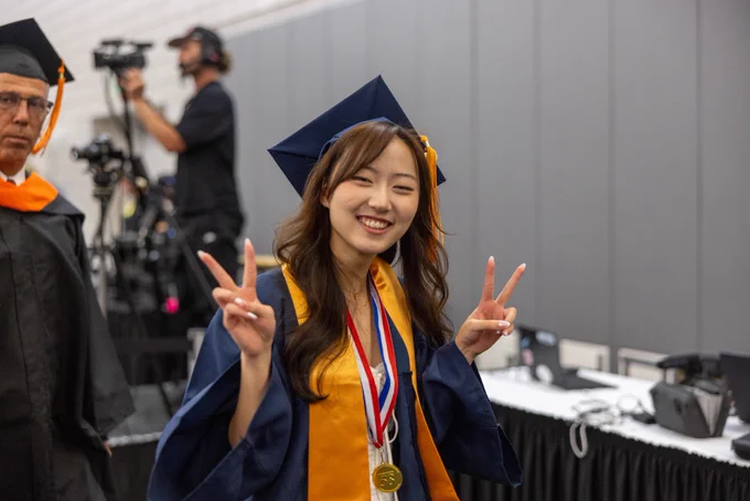 Graduate wearing a navy cap and gown with a gold stole poses indoors, holding up both hands in a peace sign; medals hang around the neck and production equipment is visible in the background.