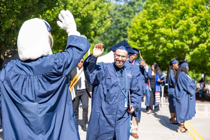 Two graduates in navy caps and gowns high-five each other outdoors along a walkway, while additional graduates in regalia gather in the background among green trees.