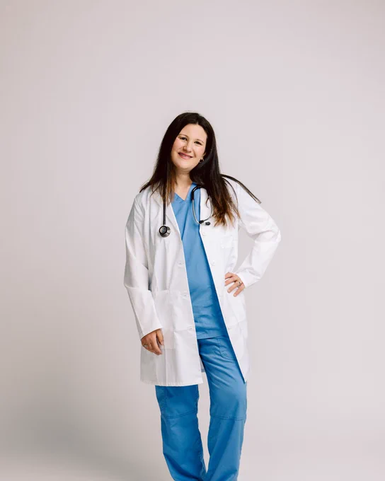 A person in blue medical scrubs and a white lab coat stands against a plain background, holding a stethoscope.
