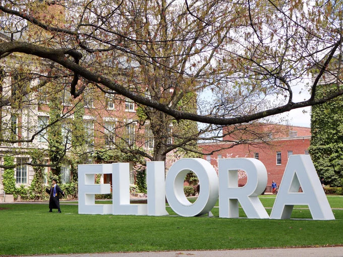 Giant white letters on the quad spell out MELIORA, with the M missing, and a student in a cap and gown stands where the missing M would be. 