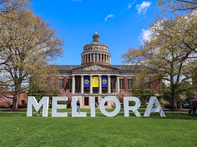 A student stands on the letter L in MELIORA in front of Rush Rhees Library.