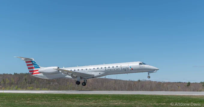 The image shows an American Eagle airplane taking off from a runway, with a clear blue sky and trees in the background.