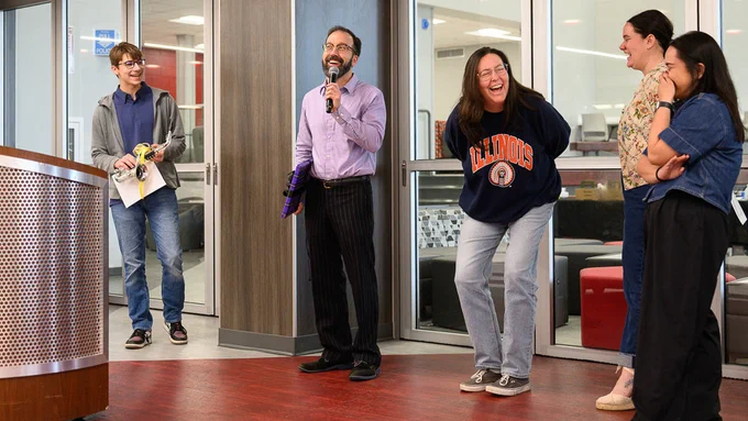 Three teachers laugh while one speaks into mic. Student smiles with award.